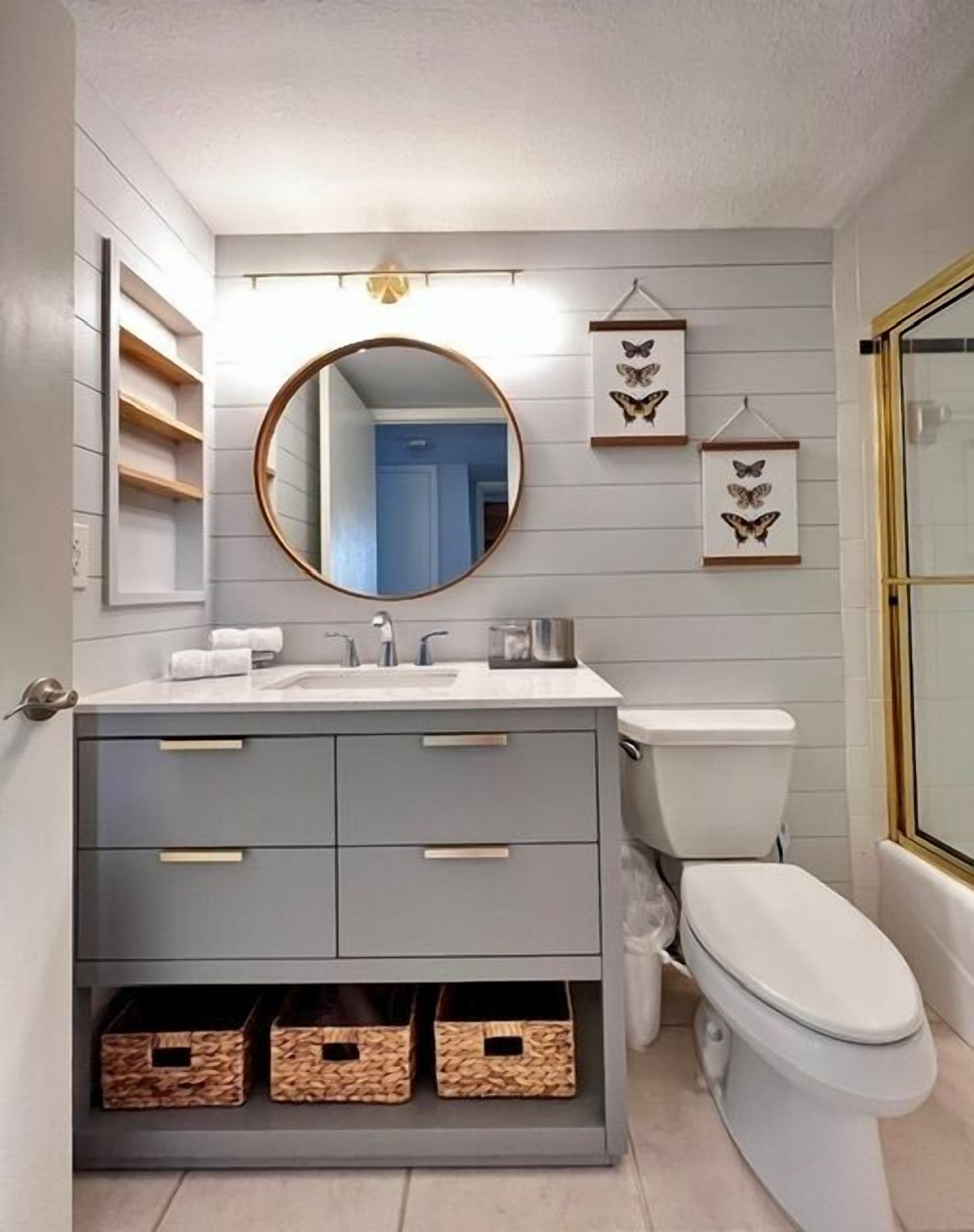 Een badkamer met een grijze wastafel, een ronde spiegel, een toilet en een wanddecoratie met vlinders.