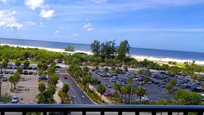 A parking lot with multiple cars and palm trees, a road leading to a beach, and a hotel building in the background. - Our House at the Beach W-701 Best Panoramic Views! (Siesta Key)
