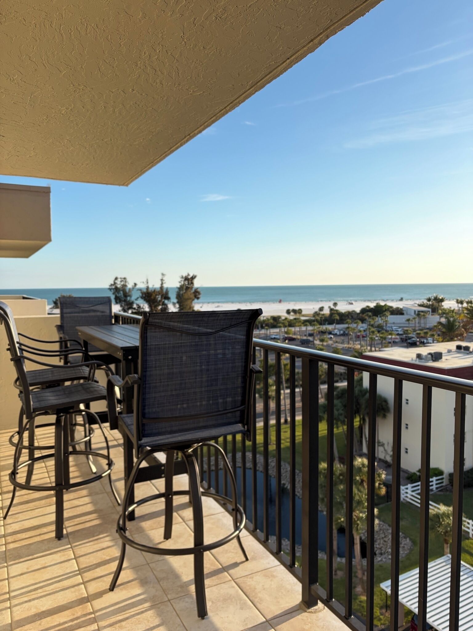 A balcony with outdoor furniture, a view of the beach, and a parking area in the background.
