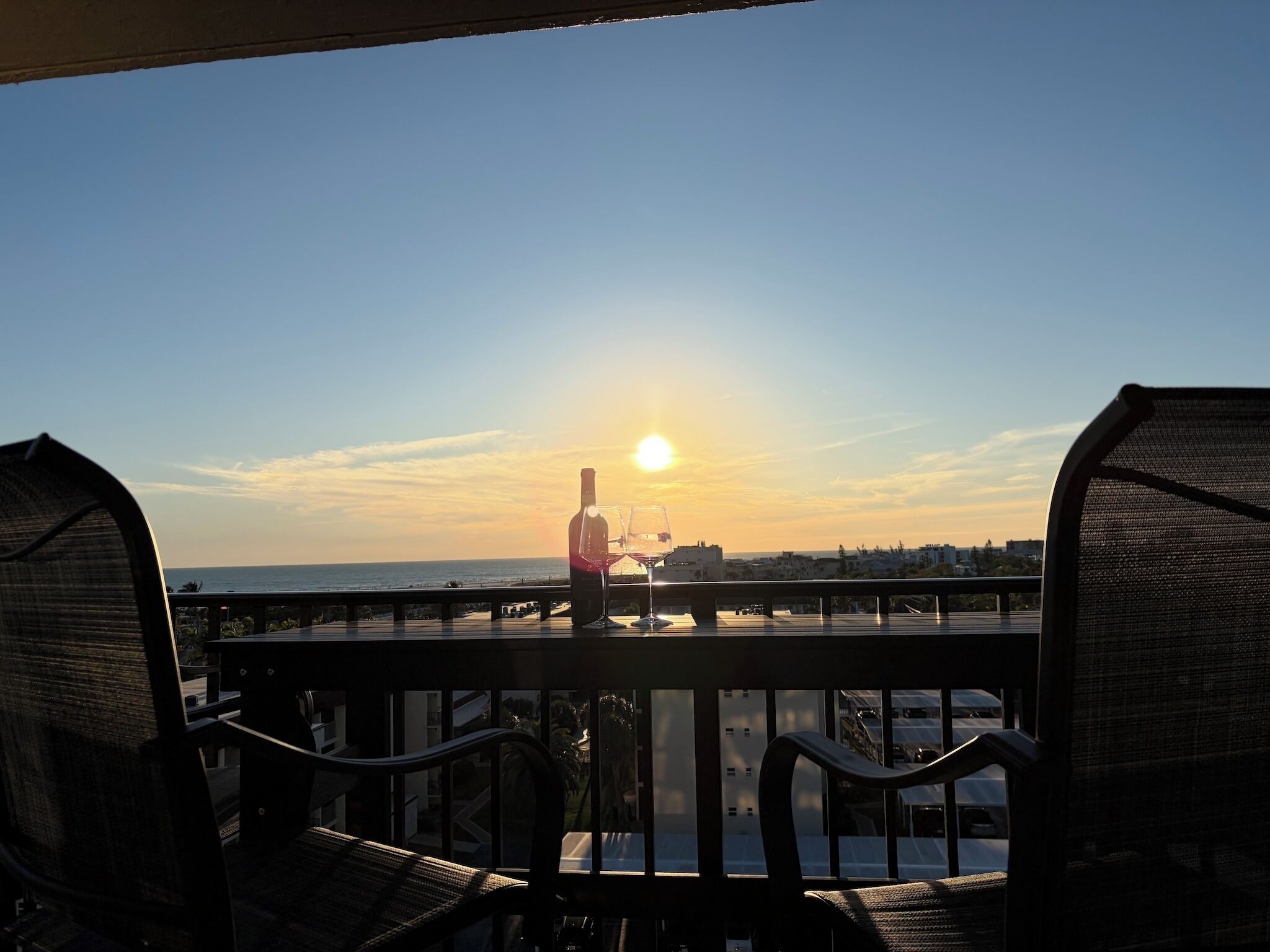 Un balcón con sillas, una botella de vino y una copa, con vistas a la puesta de sol sobre el océano.