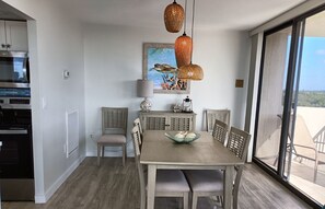 A dining area with a large table, chairs, and a bowl of fruit. There is a kitchen area with a refrigerator and oven. A large window provides a view of the outdoors.