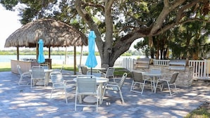 Patio esterno con ombrellone di paglia, sedie in metallo bianco e zona barbecue.