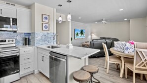 Una cocina moderna con armarios blancos, horno de acero inoxidable y lavavajillas. Hay una zona de comedor con una mesa y sillas, y un ventilador de techo.