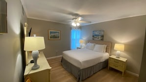 Ein Schlafzimmer mit einem Bett, zwei Nachttischen mit Lampen, einem Deckenventilator, einem Fenster mit Vorhängen und zwei Wandbildern.