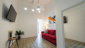 A hallway with patterned flooring, a red sofa, a ceiling fan, a TV mounted on the wall, and potted plants.
