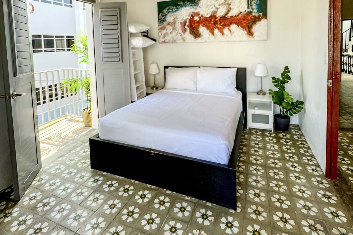 A bedroom with a bed, a nightstand, a lamp, a potted plant, and a window with a view of the outside.