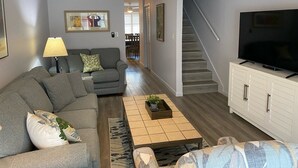 A living room with a grey sofa, a coffee table, a TV on a cabinet, and a staircase. - Townhouse with Beach Access, Shared Pool - Siesta Beach House 308 (Siesta Key)