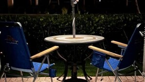 An outdoor patio with a table and two blue folding chairs, a blue umbrella, and a hedge in the background. - Sunset Royale - 100- Outdoor Patio directly across from Siesta Key Beach (Siesta Key)