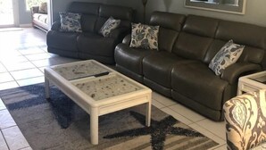 A living room with a brown leather sofa, a glass coffee table, a patterned rug, a ceiling fan, and a large mirror on the wall. - Sunset Royale - 100- Outdoor Patio directly across from Siesta Key Beach (Siesta Key)