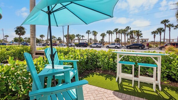A patio area with turquoise chairs and a table under a large umbrella, surrounded by lush greenery and palm trees. - Sunset Royale - 106- Directly Across from Siesta Key Beach! (Siesta Key)