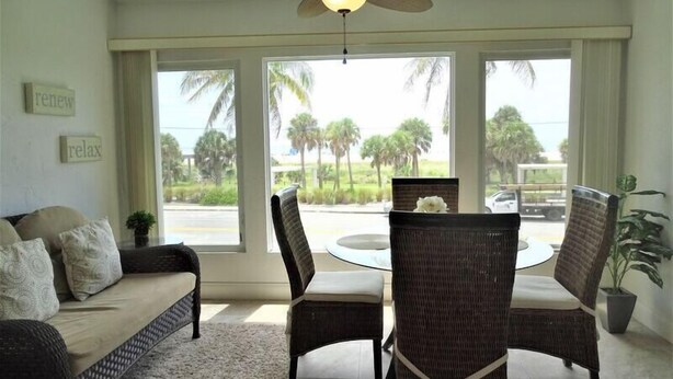 A living room with a sofa, two wicker chairs, a coffee table, and a ceiling fan. There are large windows with a view of palm trees and a road. - Sunset Royale - 200- Beautiful two bedroom, two bathroom condo with great views. (Siesta Key)