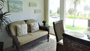 A cozy living room with a wicker sofa, a dining table, and a view of a street with palm trees. - Sunset Royale - 200- Beautiful two bedroom, two bathroom condo with great views. (Siesta Key)