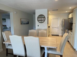 A dining area with a wooden table and four upholstered chairs, a clock on the wall, and a kitchen with white cabinets and appliances.