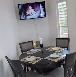 A dining area with a round table, four chairs, a television mounted on the wall, and a window with blinds.