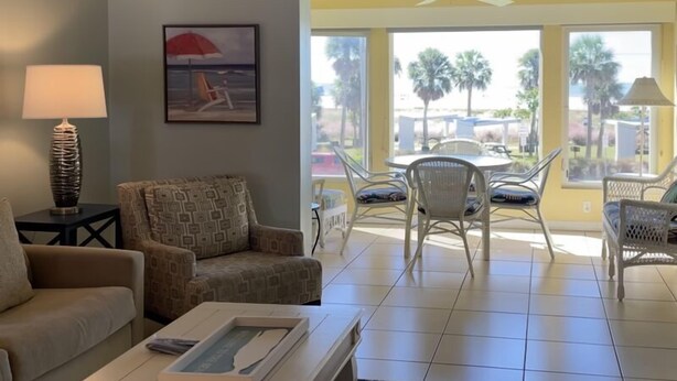 Un salon comprenant un canapé, un fauteuil et une table basse. Un tableau est accroché au mur, et une fenêtre offre une vue sur des palmiers.