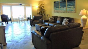 Ein Wohnzimmer mit einer Couch, Sesseln, einem Esstisch und Stühlen. An der Wand hängt ein Gemälde und ein Lampenschirm steht daneben.