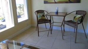 A dining area with two wicker chairs, a glass table, and a painting of palm trees on the wall. - Sunset Royale - 302- Stunning Sunset Views (Siesta Key)