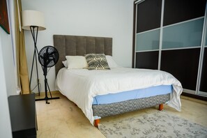 A bedroom with a bed, a standing fan, a lamp, and a large window.