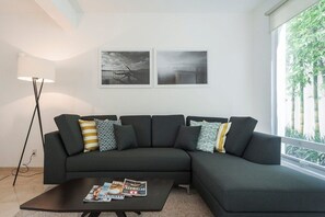 A modern living room with a dark grey sectional sofa, a coffee table with magazines, a floor lamp, and two framed pictures on the wall.