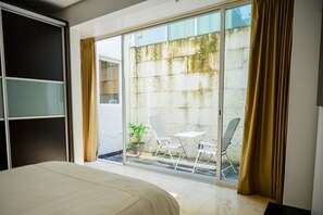 A hotel room with a bed, a sliding glass door leading to a balcony with a table and chairs, and a potted plant.
