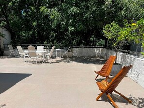 An outdoor seating area with lounge chairs and tables, surrounded by greenery and a concrete wall.