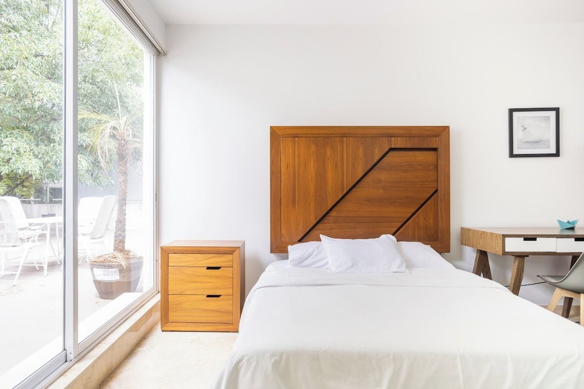Una camera da letto con un letto, un comodino in legno, una scrivania e una finestra vista su sedie da esterno e una palma.