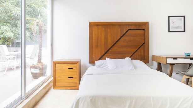 A bedroom with a bed, a wooden nightstand, a desk, and a window with a view of outdoor chairs and a palm tree. - Condesa Residence Amsterdam G (CDMX)