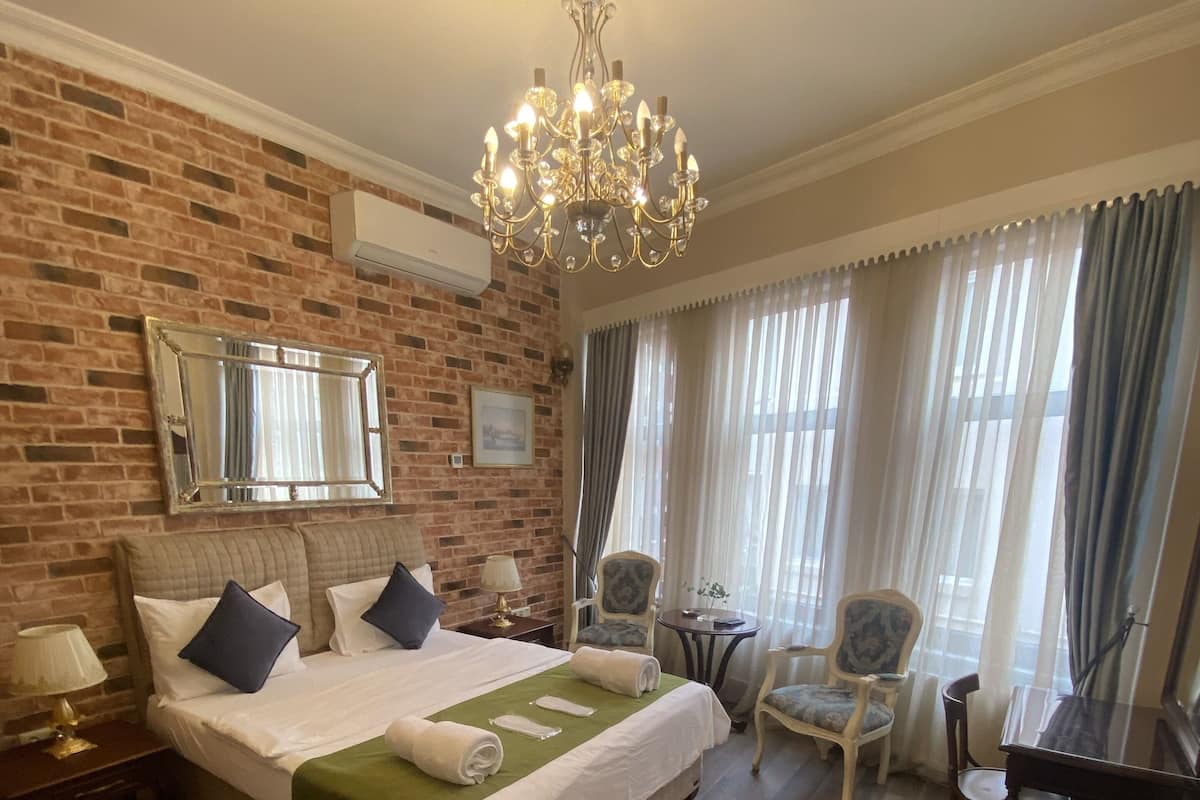 A hotel room with a brick wall, a chandelier, a bed with a green bedspread, a wooden floor, a dining table, and a chair.
