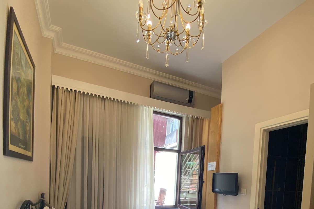 A hotel room with a bed, a chair, a table with a plant, a chandelier, a window with curtains, and a door leading to a bathroom.
