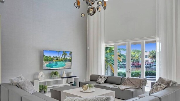 A modern living room with a large window, a flat-screen TV, and a view of a pool and greenery. - On The Bay With Hot Tub, Pool, Kayaks And More (Hollywood)