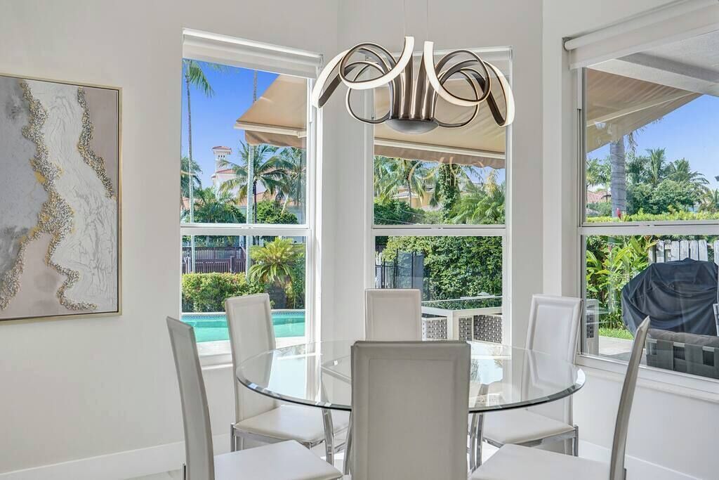 Un espace repas avec une table en verre et des chaises blanches, de grandes fenêtres offrant une vue sur les palmiers et une piscine, ainsi qu’un luminaire moderne.