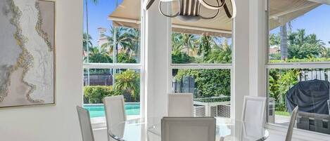 Un espace repas avec une table en verre et des chaises blanches, de grandes fenêtres offrant une vue sur les palmiers et une piscine, ainsi qu’un luminaire moderne.