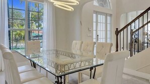 A dining area with a glass table and white upholstered chairs, a chandelier, and a staircase with a wrought iron railing. - On The Bay With Hot Tub, Pool, Kayaks And More (Hollywood)