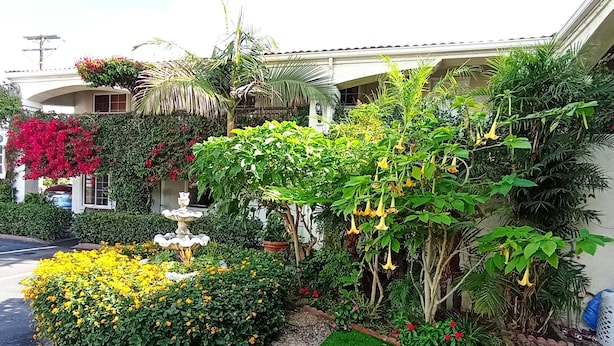 Um jardim bem cuidado com uma fonte, diversas plantas floridas e um gramado aparado em frente a um edifício com telhado curvo e varanda.