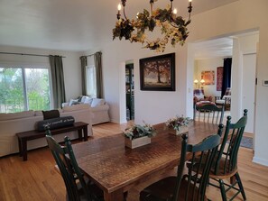 Un comedor con una mesa y sillas de madera, un sofá con cojines, un cuadro en la pared y vistas al exterior a través de una ventana.