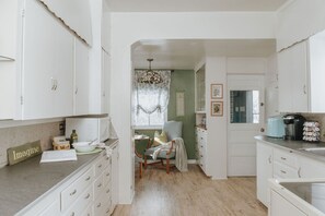 A kitchen with white cabinets, a dining area with a chair, a window with lace curtains, and a sign that says "Imagine". - Amor Vida — a quiet 1910 farmhouse in the Colorado countryside (Ault)