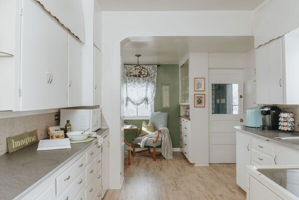 A kitchen with white cabinets, a dining area with a chair, a window with lace curtains, and a sign that says "Imagine". - Amor Vida — a quiet 1910 farmhouse in the Colorado countryside (Ault)