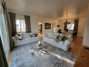 A spacious living room with a large sectional sofa, a glass coffee table, and a dining area with a wooden table and chairs. - Amor Vida — a quiet 1910 farmhouse in the Colorado countryside (Ault)