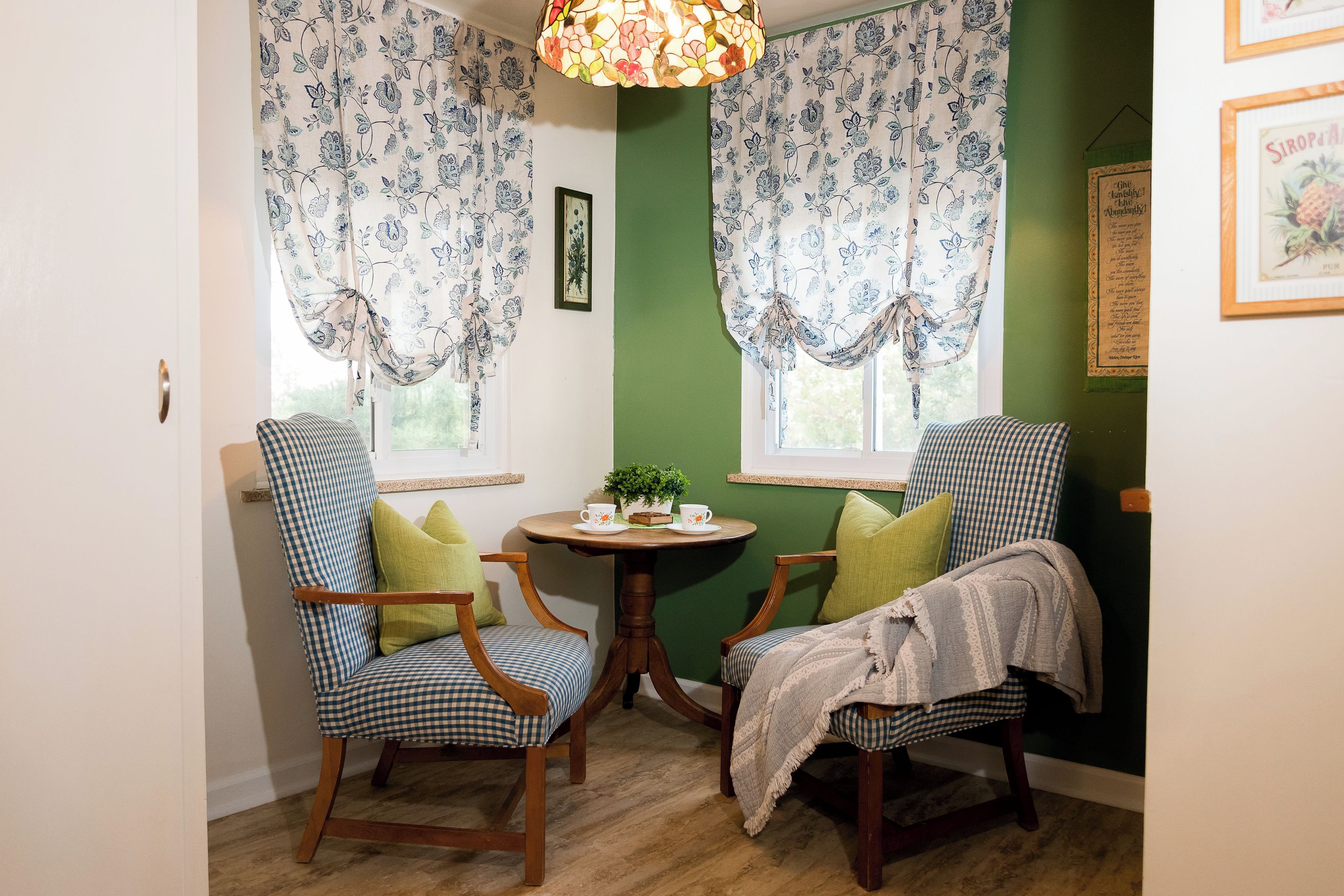 A room with two checkered chairs, a small round table with a cup and saucer, and floral curtains.