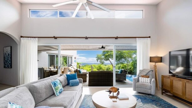A spacious living room with a large sofa, a round coffee table, and a television. There is a sliding glass door leading to an outdoor area with a pool and furniture. - Amigos de Tortuga • 4 BR Villa • Private Pool • Oceanview (Playa Hermosa)