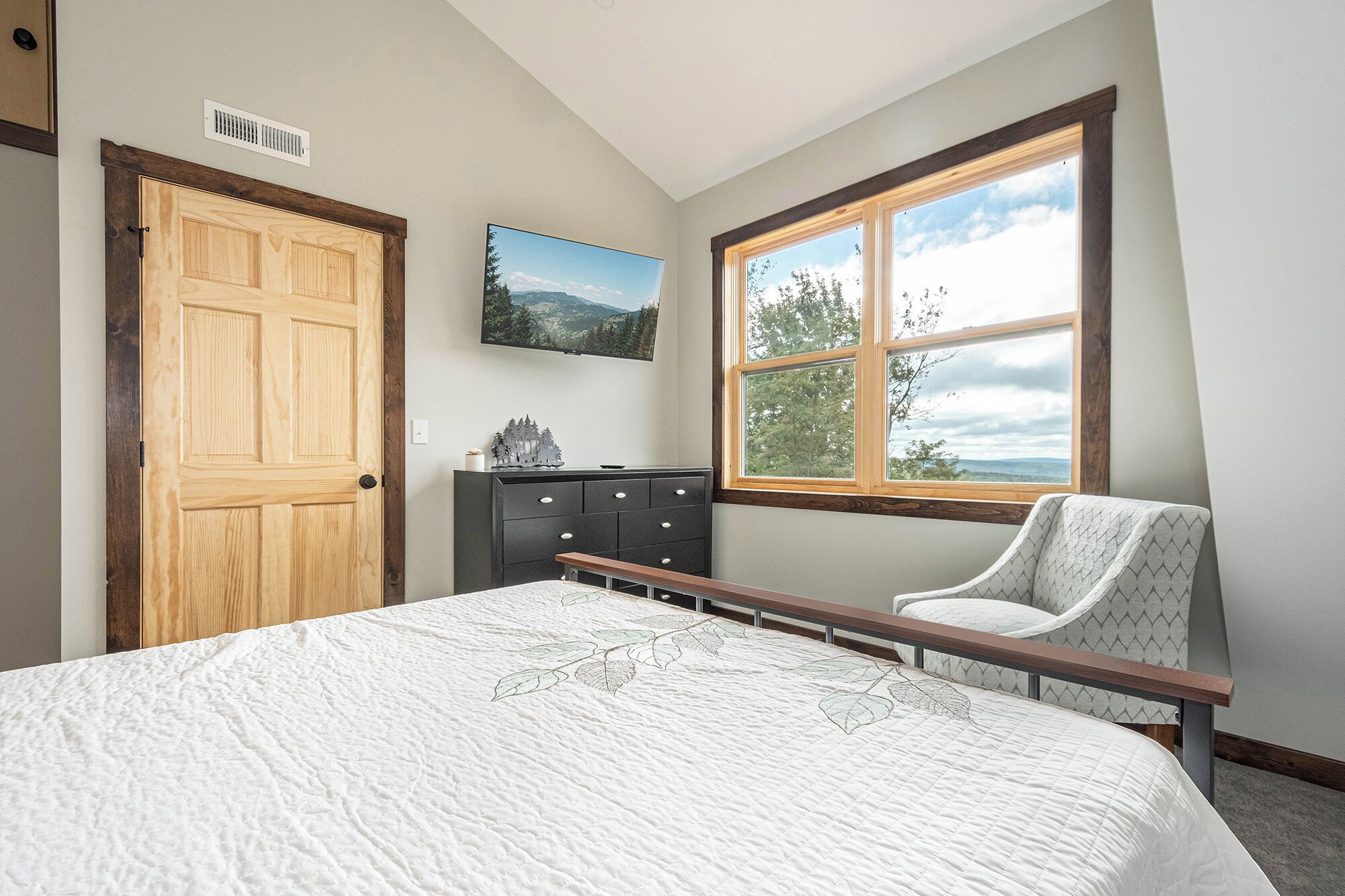 A bedroom with a large bed, a dresser, a chair, a window with a view of trees, and a door.
