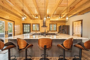 A modern kitchen with a large island, bar stools, stainless steel appliances, and wooden ceiling beams. - Sunshine Chalet by Taylor Made Deep Creek (McHenry)