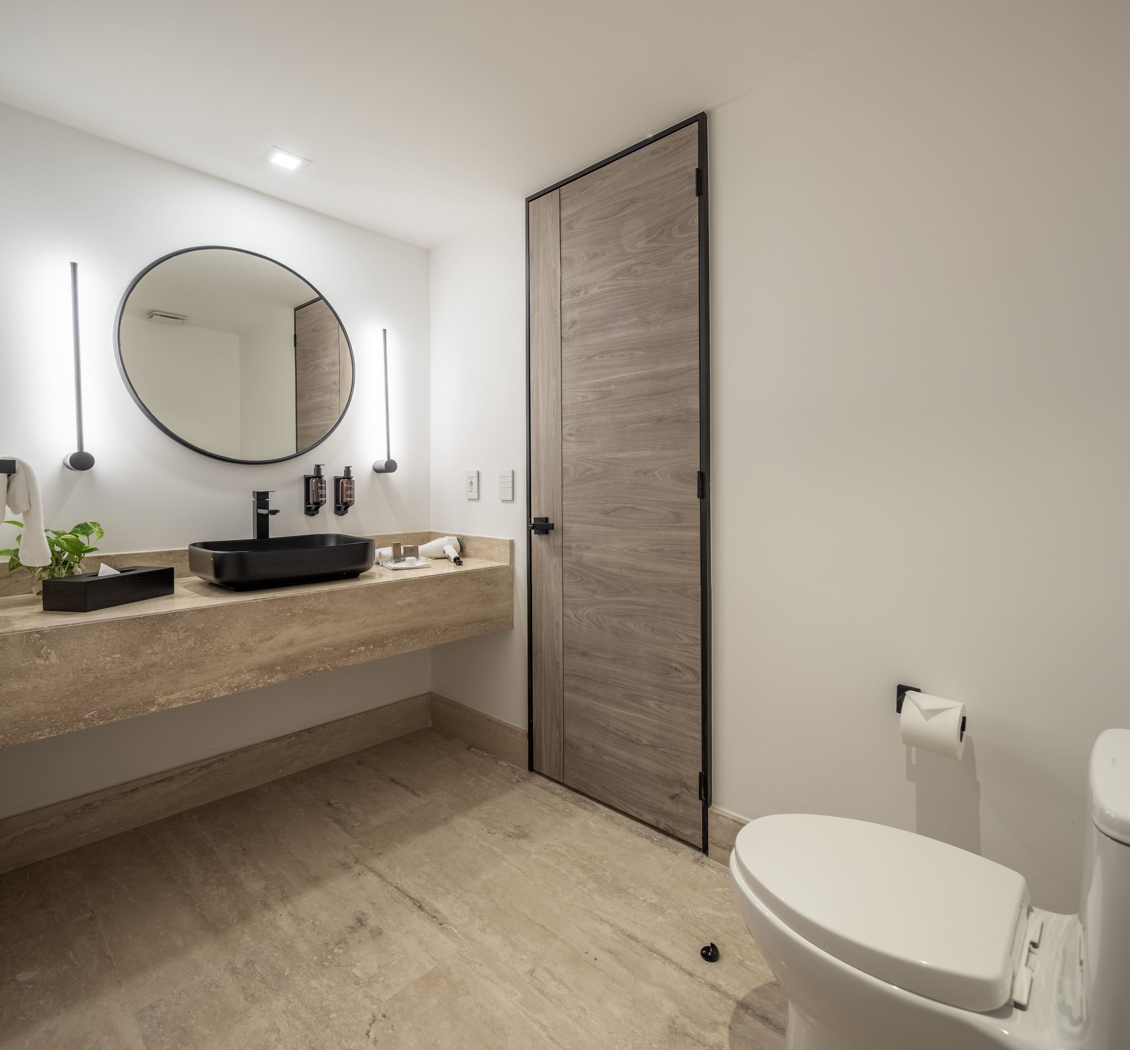 Un baño moderno con un espejo redondo, un lavamanos negro, un inodoro y una puerta de madera.