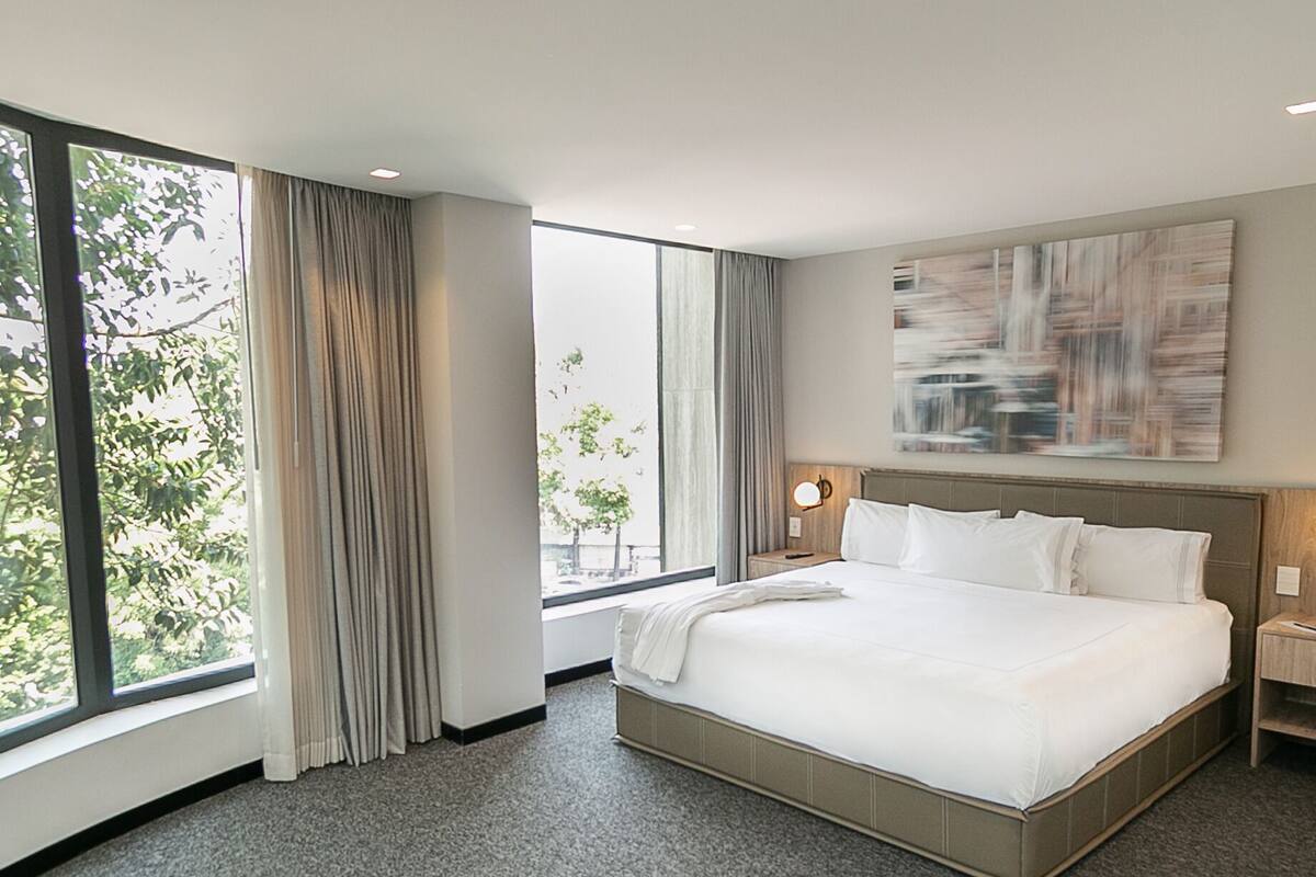 A modern hotel room with a large bed, a view of trees through the window, and a painting on the wall.