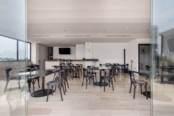 Un comedor moderno con varias mesas y sillas, una zona de cocina con televisor de pantalla plana y vistas a través de un panel de vidrio.