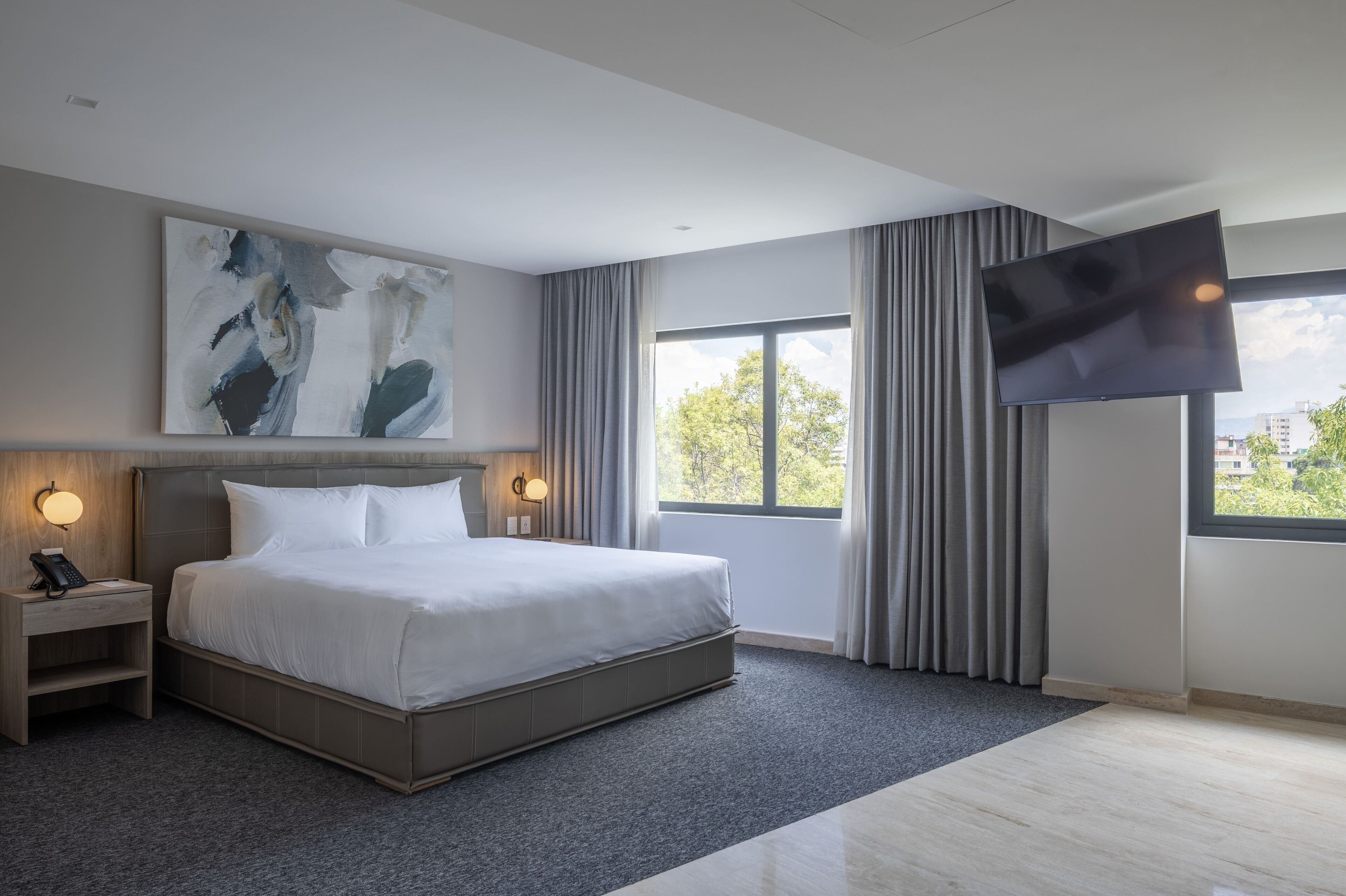 A modern hotel room with a large bed, a flat-screen TV, a painting on the wall, and a view of the outdoors through the window.