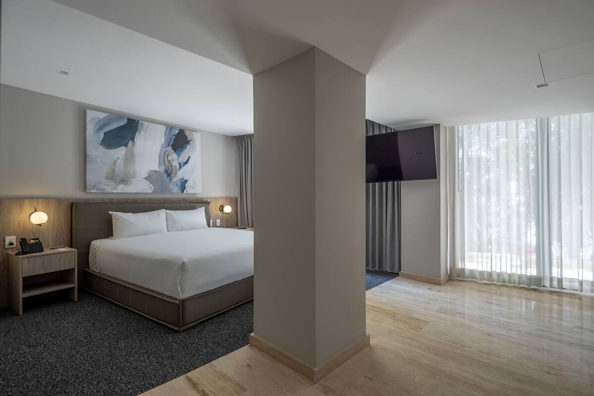 A modern hotel room with a large bed, a flat-screen TV, a painting on the wall, and a large window with curtains.