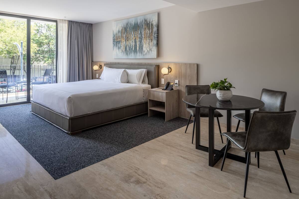 A modern hotel room with a large bed, a desk with a telephone, and a dining area with chairs and a table.