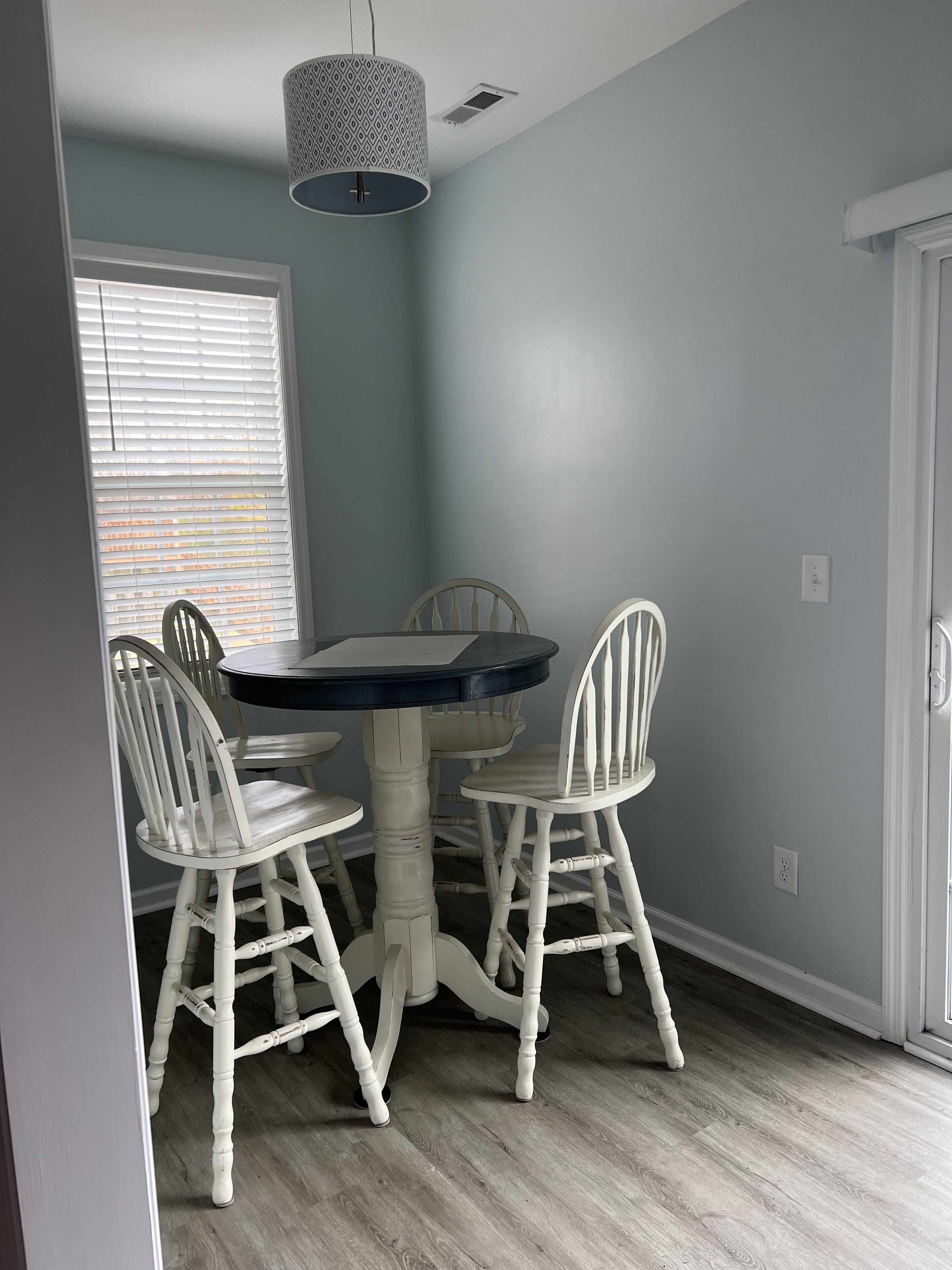 A dining area with a round table and four white chairs, a hanging light fixture, and a window with blinds.