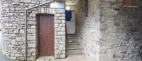 A stone building with a wooden door, a metal pipe, and a staircase leading to an upper level.